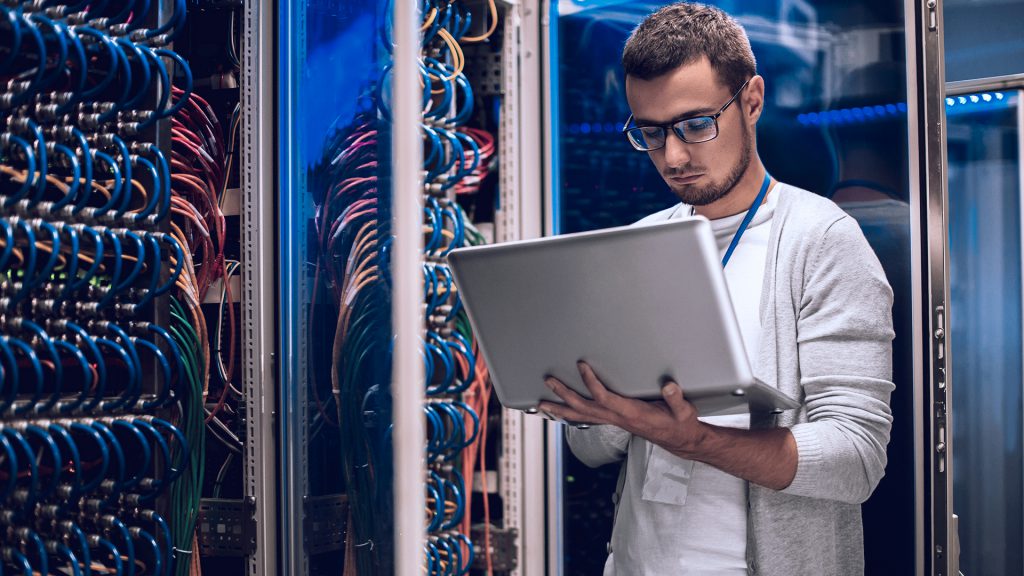 Foto de analista de T.I. segurando notebook, em frente a um data center.