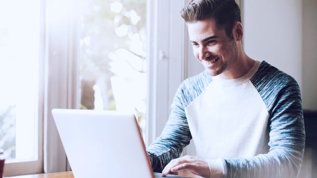 Foto de homem com expressão feliz enquanto usa um notebook em casa.